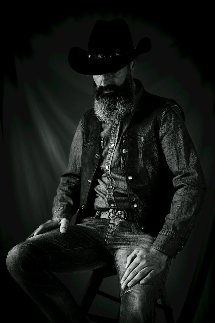services-02 A dramatic black and white portrait of a bearded cowboy wearing a hat and denim clothing, seated in a studio.